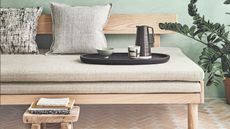 A sage green room with Japanese futon sofa with cushions, tray with teapot and cups, and a wooden stool with books. The tiles are pale terracotta, with a large houseplant to the right.