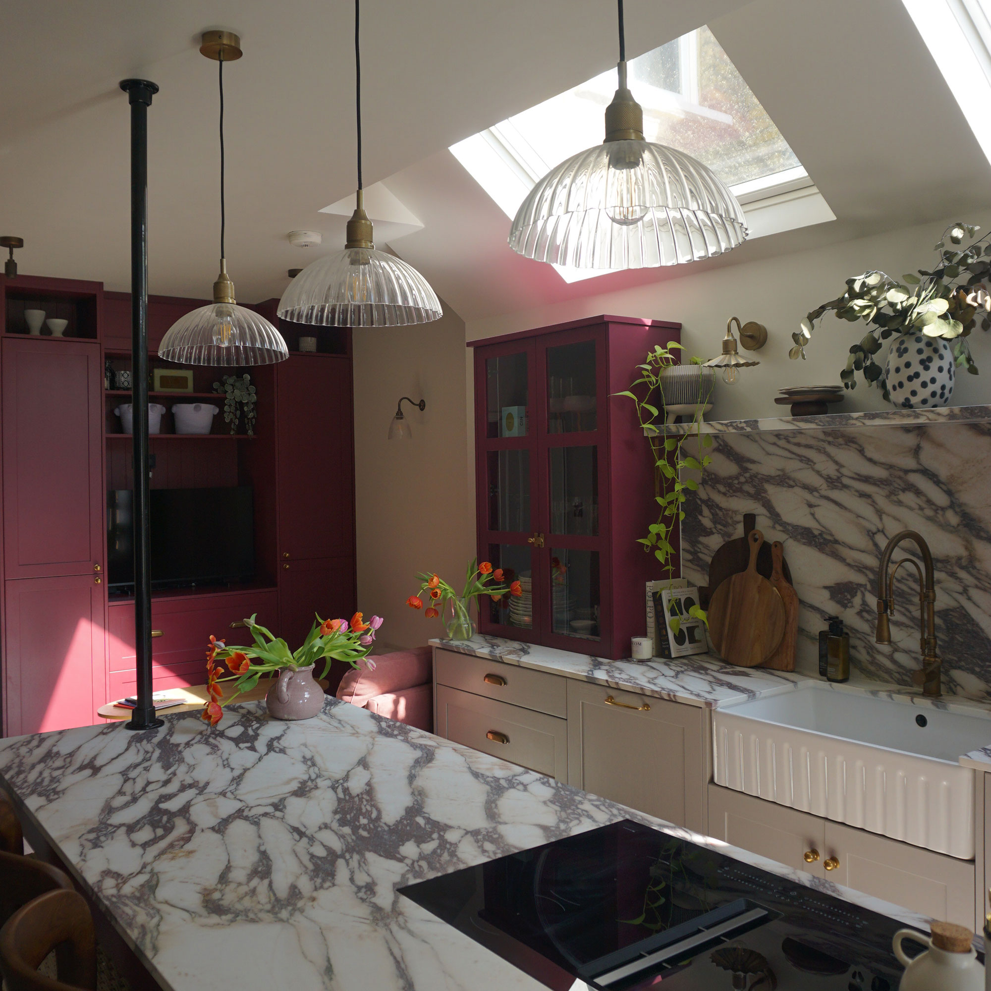 Marble kitchen worktop run with deep fluted Belfast sink, marble splashback and red cabinetry across from marble topped island with integrated hob