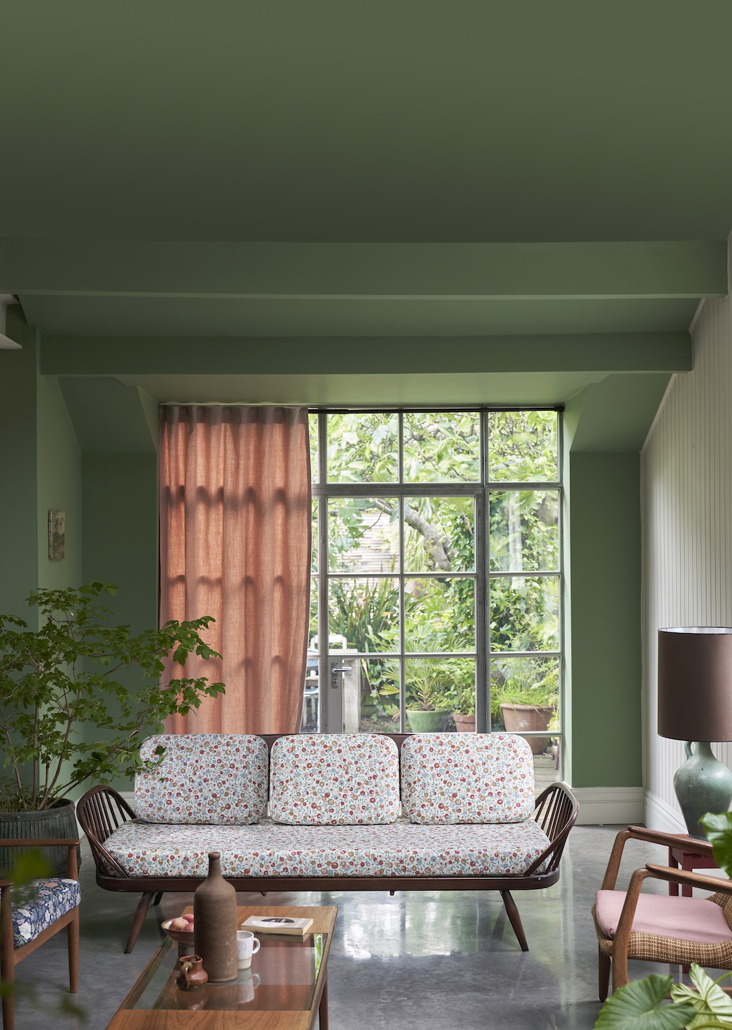 Green Farrow &amp;amp; Ball paint in an earthy living room that has large windows to a back garden. There is a white, dainty floral sofa.