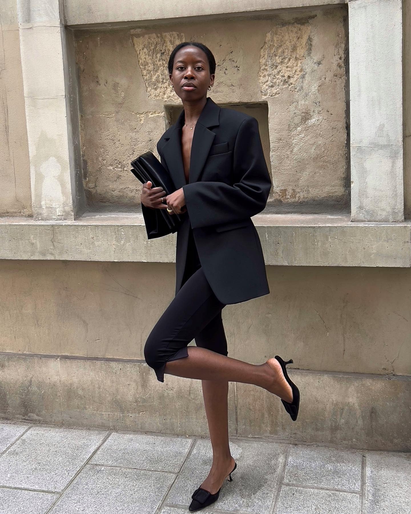Paris-based fashion influencer and stylist Sylvie Mus poses on a sidewalk wearing an oversize black blazer, black leather bag, black capri pants, and Manolo Blahnik Maysale suede mule heels