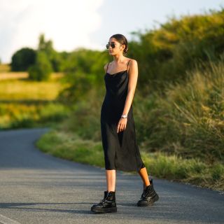 Jacqueline Barth wears sunglasses, a black lustrous silky long dress, black leather boots, on July 26, 2021 in Sibiu, Romania.