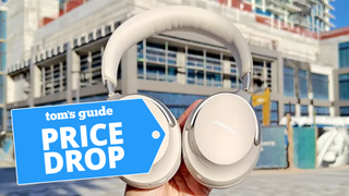 Bose QuietComfort Ultra Headphones being held up in front of urban building with price drop badge