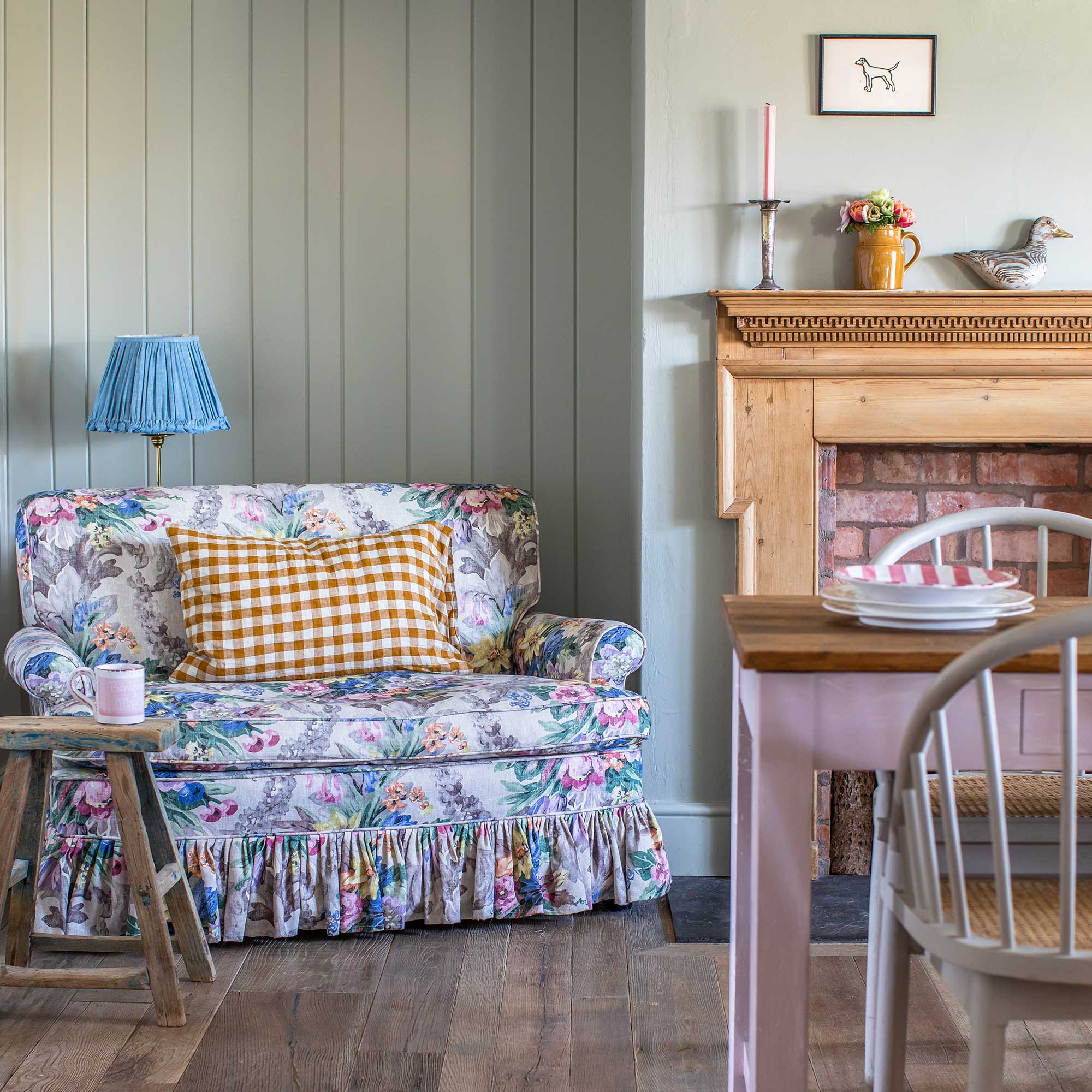 small sofa covered in floral fabric in alcove with green painted tongue and groove panelling, beside wooden fireplace and pink farmhouse table