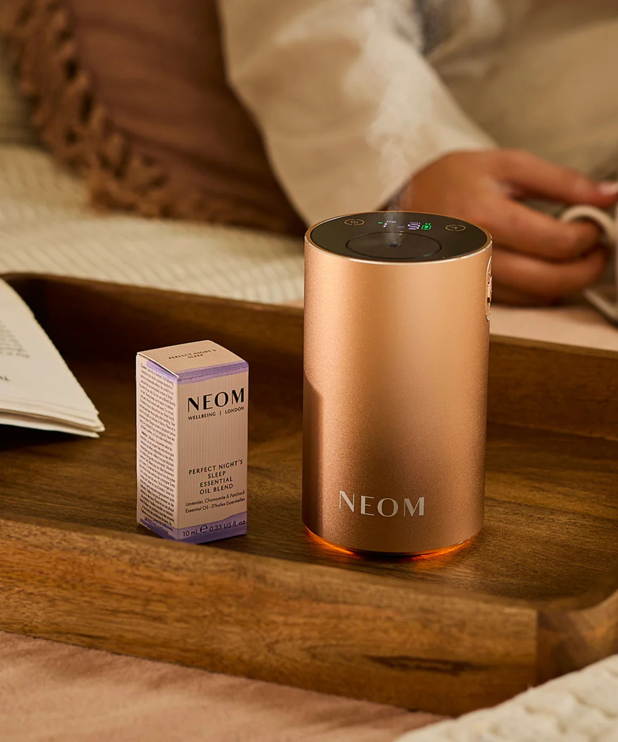A rose gold Neom Wellbeing Pod Mini+ on a wooden tray, beside a pale pink and purple cardboard packaging box of essential oil. In the background, someone is sitting holding a mug of tea.