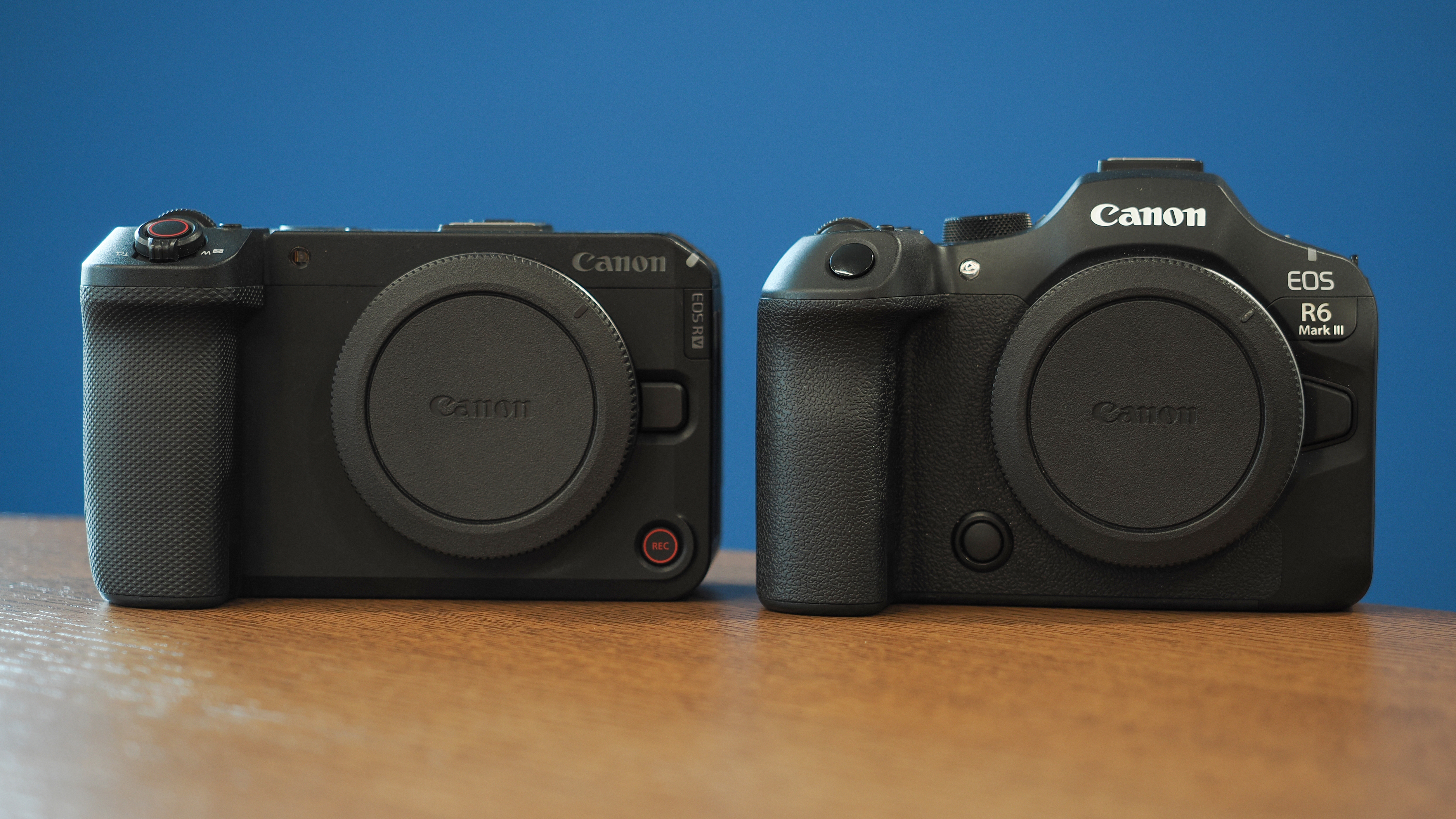 Canon EOS R6 V and R6 Mark III on a wooden surface, in front of a blue background