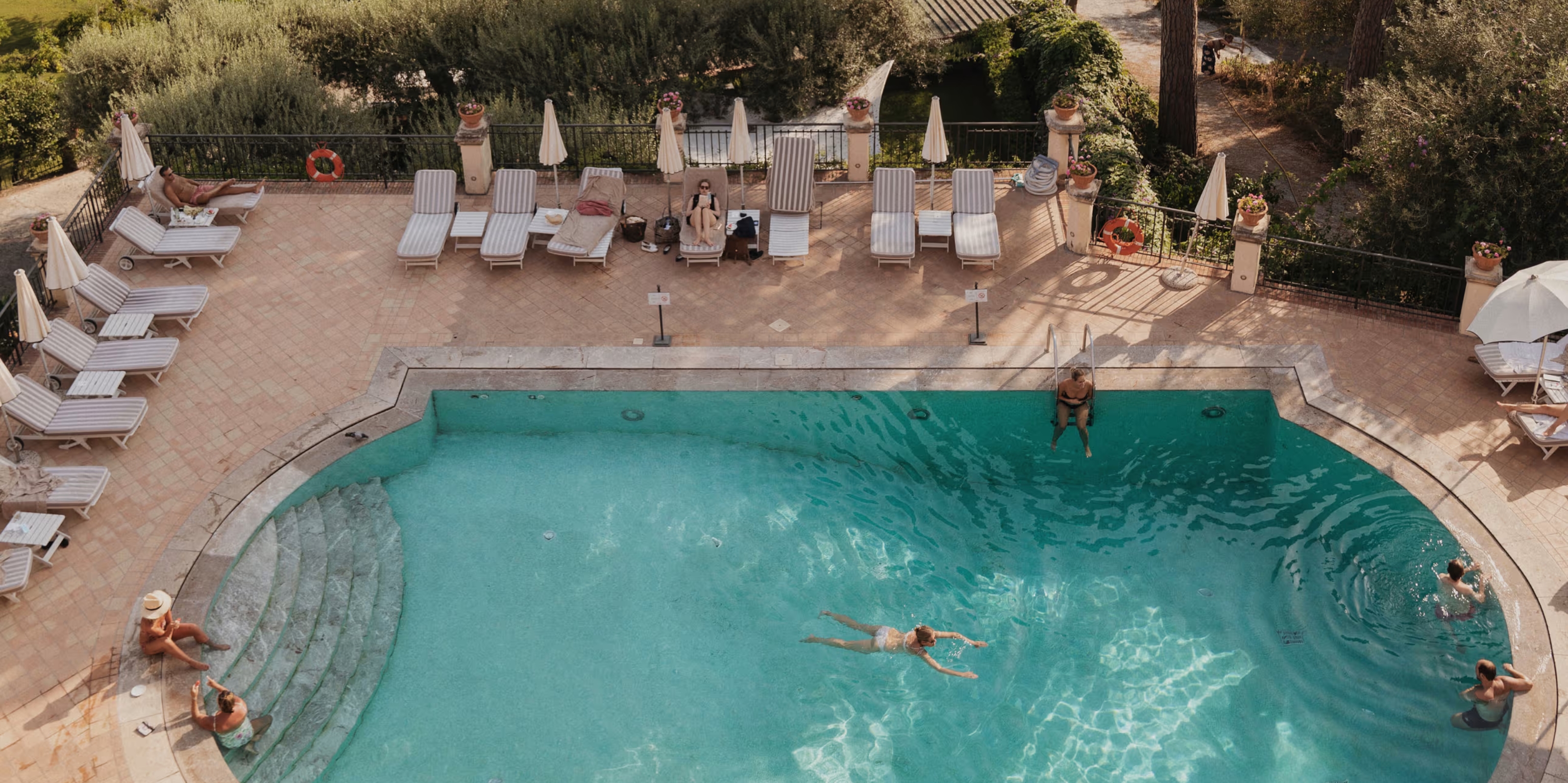 An aerial view of a resort pool shows a series of people bathing in its waters in dim afternoon sunshine, with vegetation and terracotta tiles surrounding them.