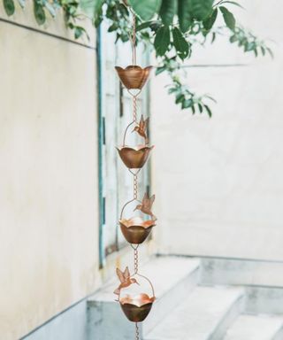A copper flower rain chain with bird cut-outs adorning it, hanging from a green leafy tree with a beige wall and gray door behind it