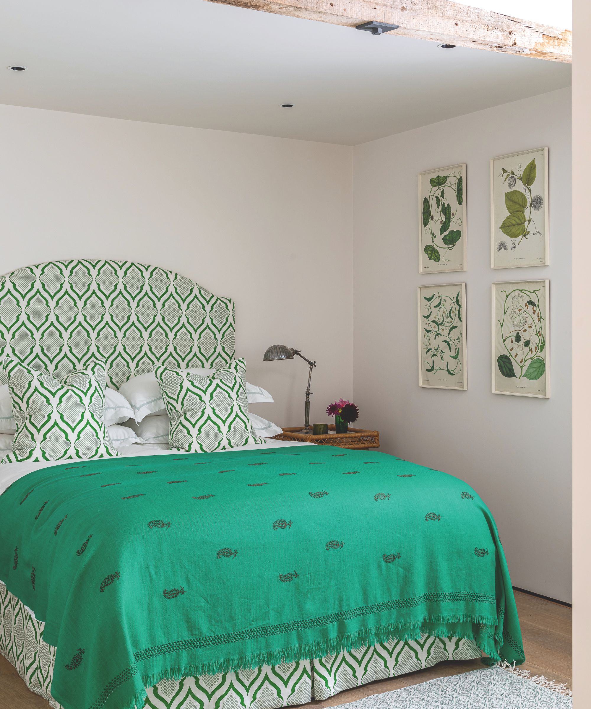 A green and white bedroom with white walls and exposed wood beams, a green and white patterned bed with matching cushions, green blanket, wooden flooring, green and white rug, wooden bedside table, and silver lamp.