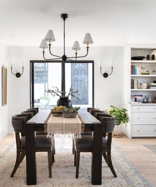 White dining room with dark wood table and chairs, vintage style rug, pale wood flooring, crittall doors with view outside, pendant light above table, two matching wall lights each side of doors, bookcase to the right