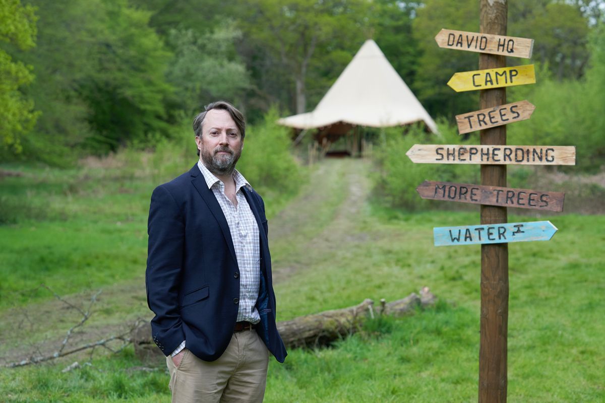 Outsiders: David Mitchell stands in the woods next to a wooden signpost with branches pointing to &#039;David HQ&#039;, &#039;Camp&#039;, &#039;Trees&#039;, &#039;Shepherding&#039;, &#039;More Trees&#039; and &#039;Water&#039;, with a tent in the background