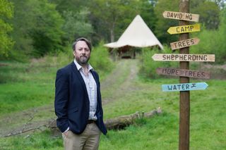 Outsiders: David Mitchell stands in the woods next to a wooden signpost with branches pointing to 'David HQ', 'Camp', 'Trees', 'Shepherding', 'More Trees' and 'Water', with a tent in the background