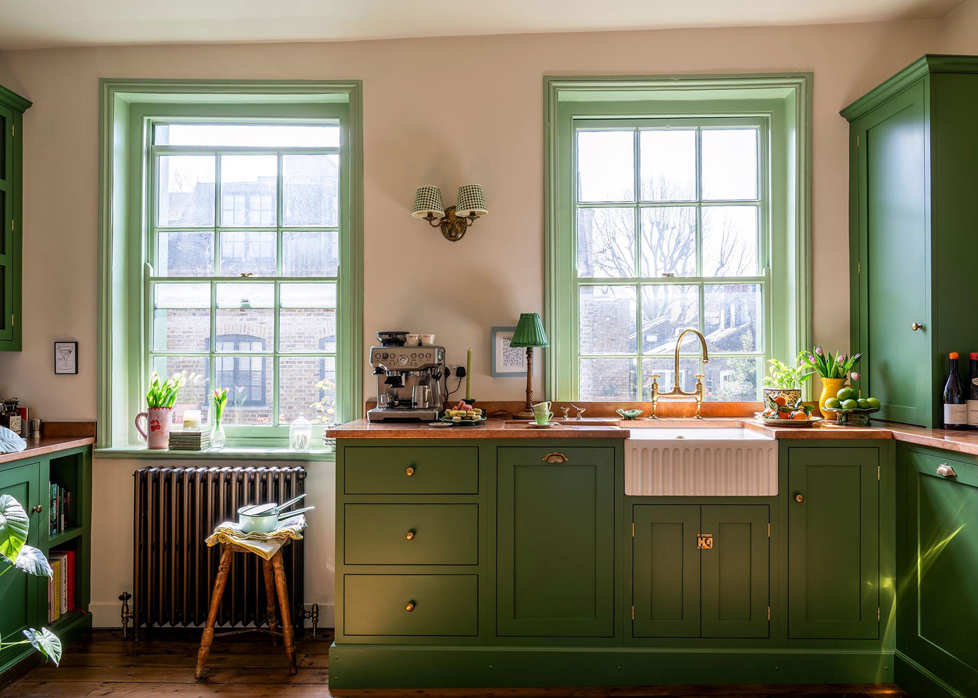green wooden kitchen with fluted ceramic sink as as well as a view or two large window and between those windows is a light fixture on the wall