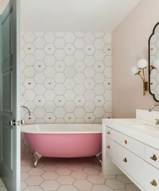 bathroom with pink painted roll top tub, large hexagon white tiles on one wall with gold bees, matching floor, white vanity, large mirror, globe lights,