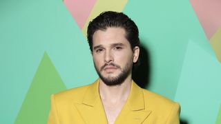 Kit Harington in a yellow jacket in front of a green background.