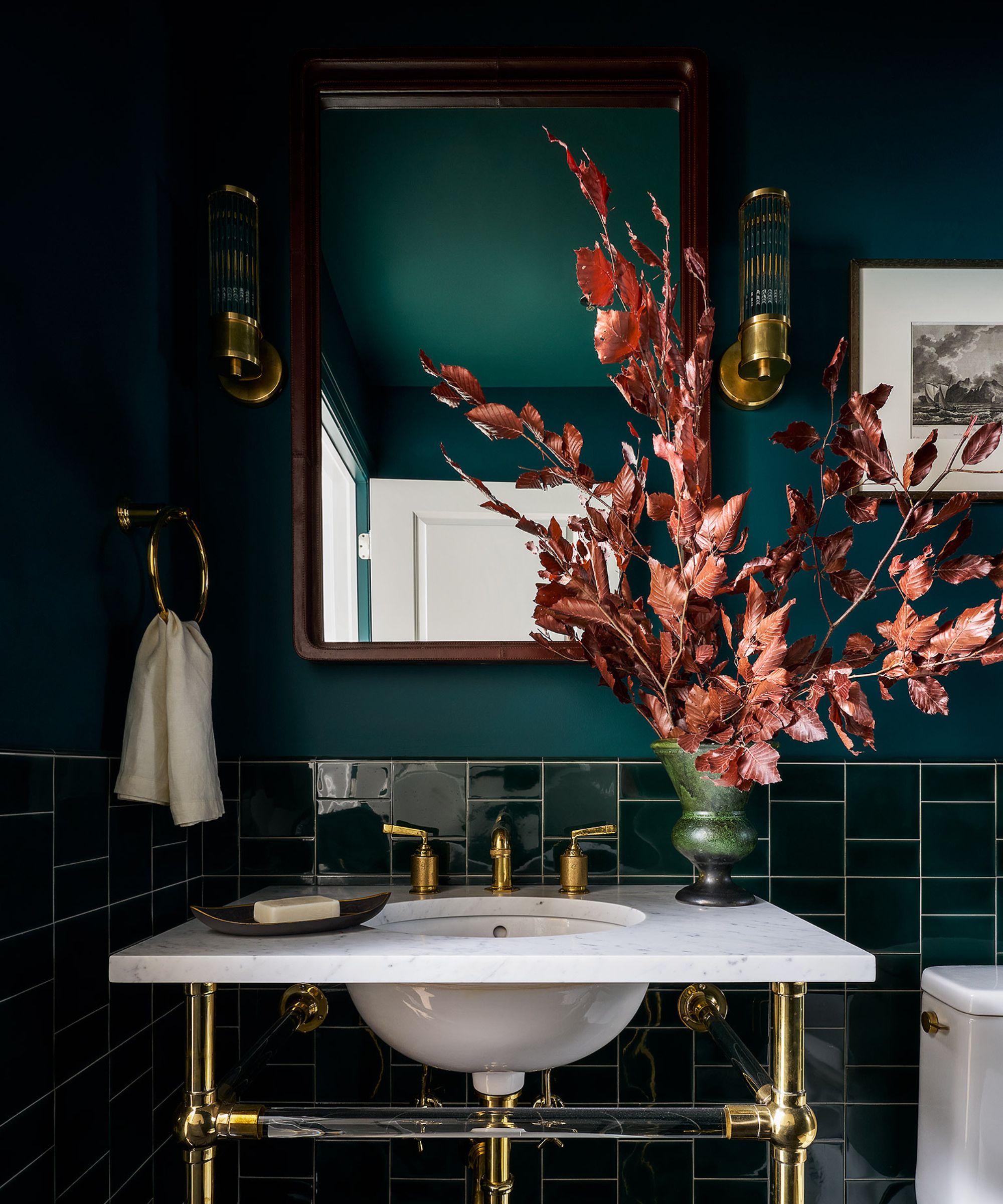 A powder room with teal walls, teal tiles on the lower wall, a white sink with brass hardware, and a vase of purple foliage.