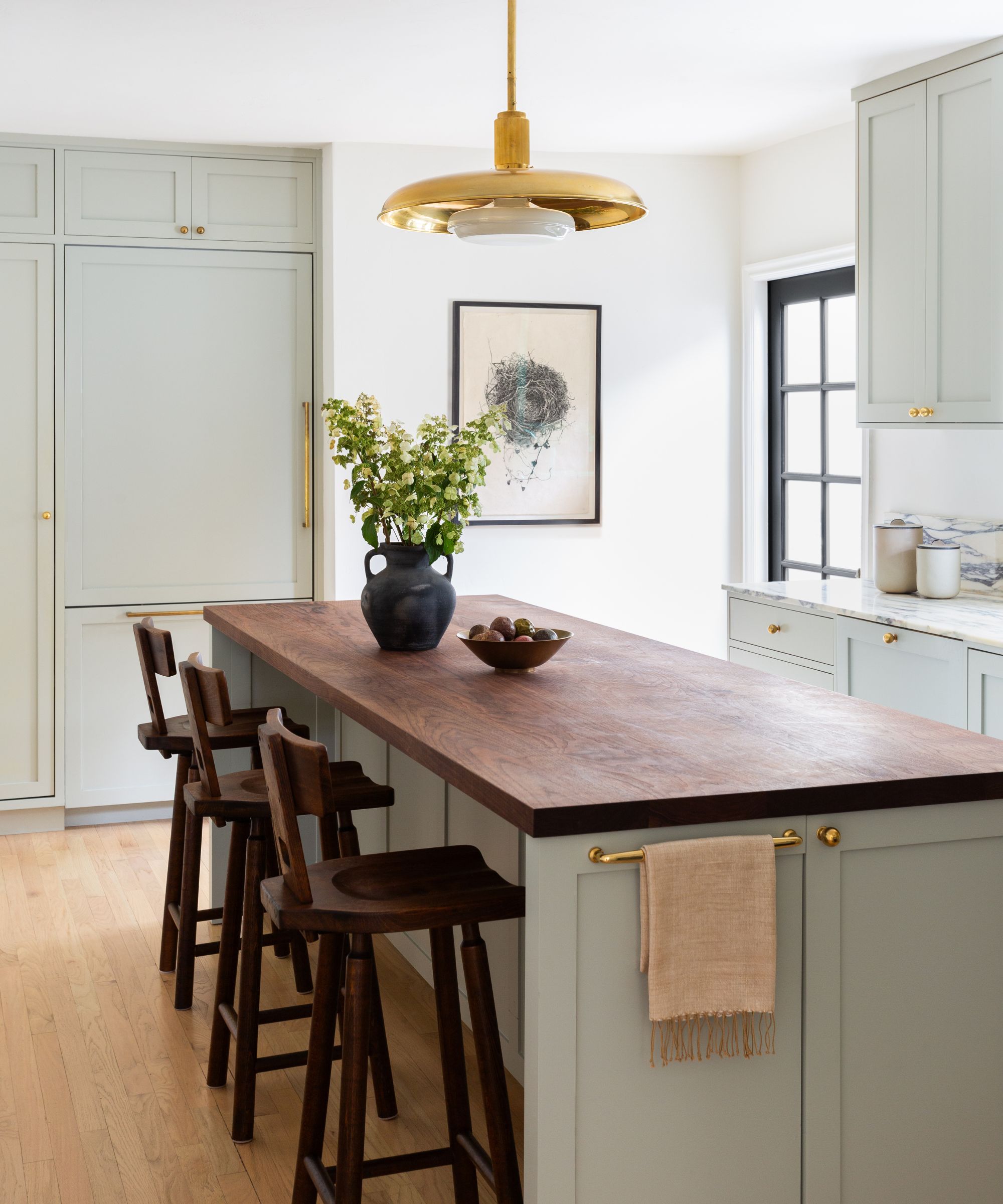 a soft green gray small kitchen with a small island with a dark wooden countertop, dark wood vintage bar stools and a brass overhead pendant