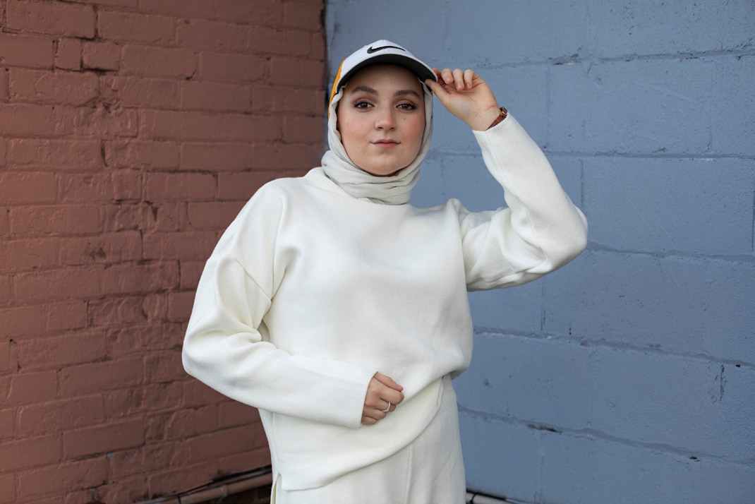 A young woman wearing a grey hijab and a white Nike cap tips the peak of her cap toward the camera, dressed in an oversized white sweatshirt, standing between a red brick wall and a blue-painted wall.