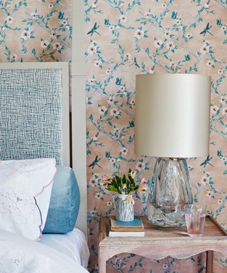 The corner of a bed; white bedding on a blue bed frame; to the right, a nightstand topped with a lamp and a vase of flowers; against a wall covered with loral beige and blue wallpaper.