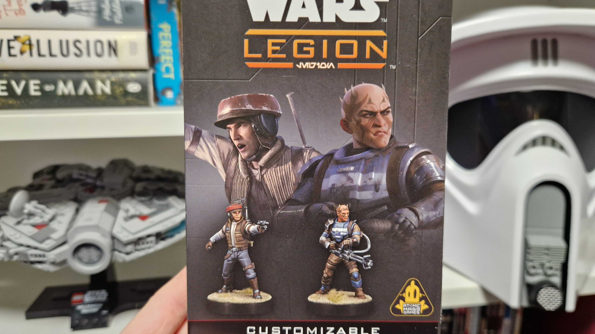 A hand holds up a box of Star Wars: Legion Rebel Alliance troopers in front of a Lego Millennium Falcon and Scout Trooper helmet