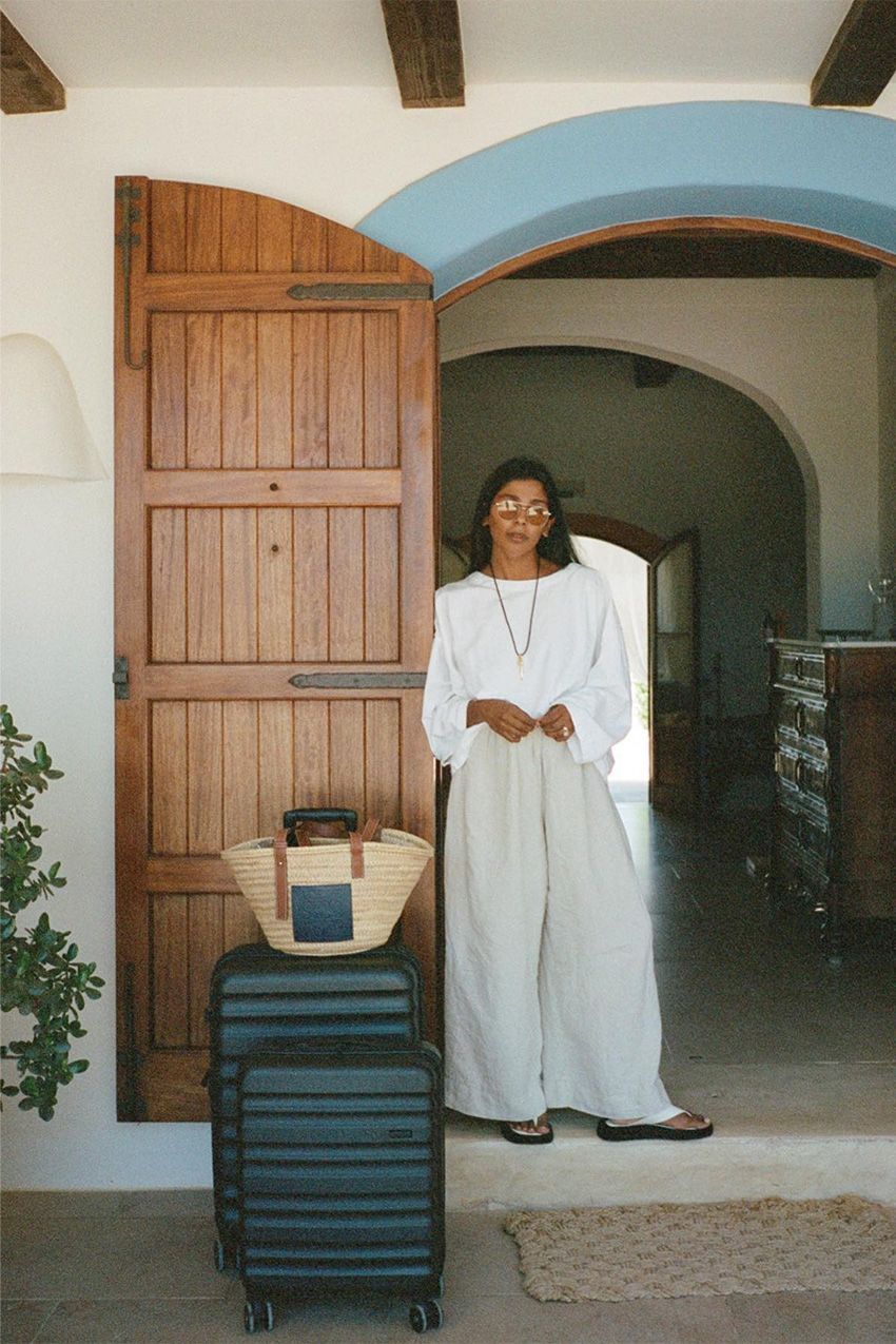European vacation outfit idea is shown in a photo of a woman standing in a doorway at a hotel by her black luggage wearing brown aviator sunglasses, an oversized white t-shirt, a long cord necklace, wide-leg tan linen trousers, with white platform flip flops and a raffia tote bag