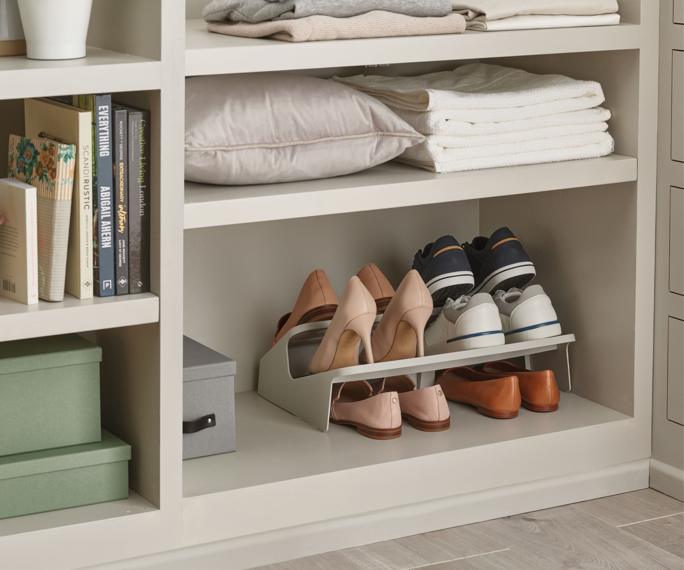 A close-up of a tiered shoe rack holding heels, trainers and flats within an open shelf of a soft grey wardrobe unit, with folded linen and a cushion on the shelf above and stacked storage boxes and books alongside.