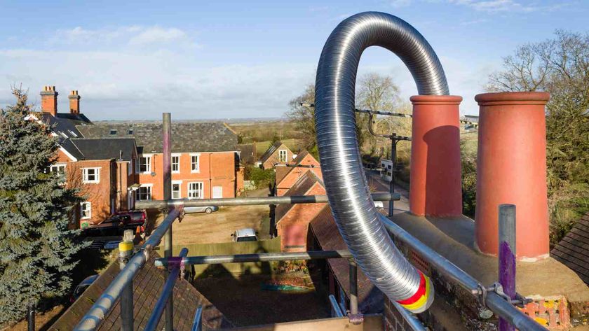 A long steel flue pipe being installed in a chimney, likely to be for a woodburning stove inside. Scaffolding surrounds the chimney and roof, and red brick houses can be seen in the background.