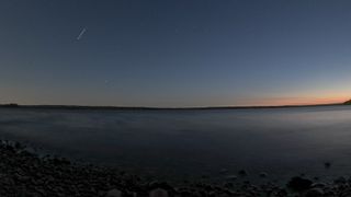 A starry night sky with the beginnings of sunrise in the right side over a lake shore