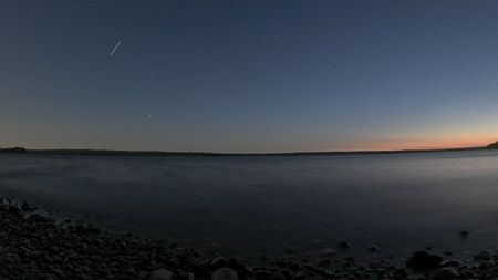 A starry night sky with the beginnings of sunrise in the right side over a lake shore