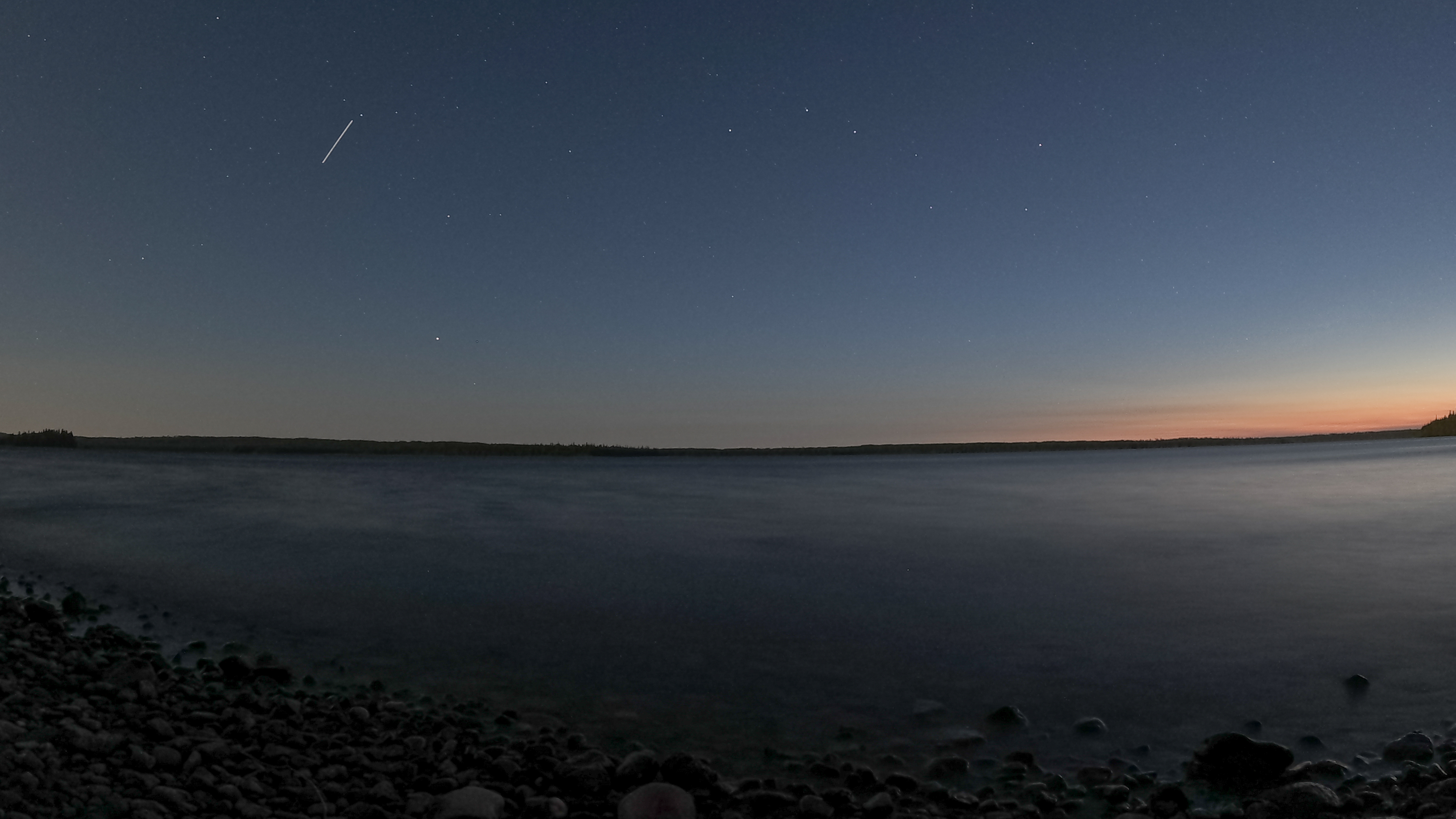 A starry night sky with the beginnings of sunrise in the right side over a lake shore