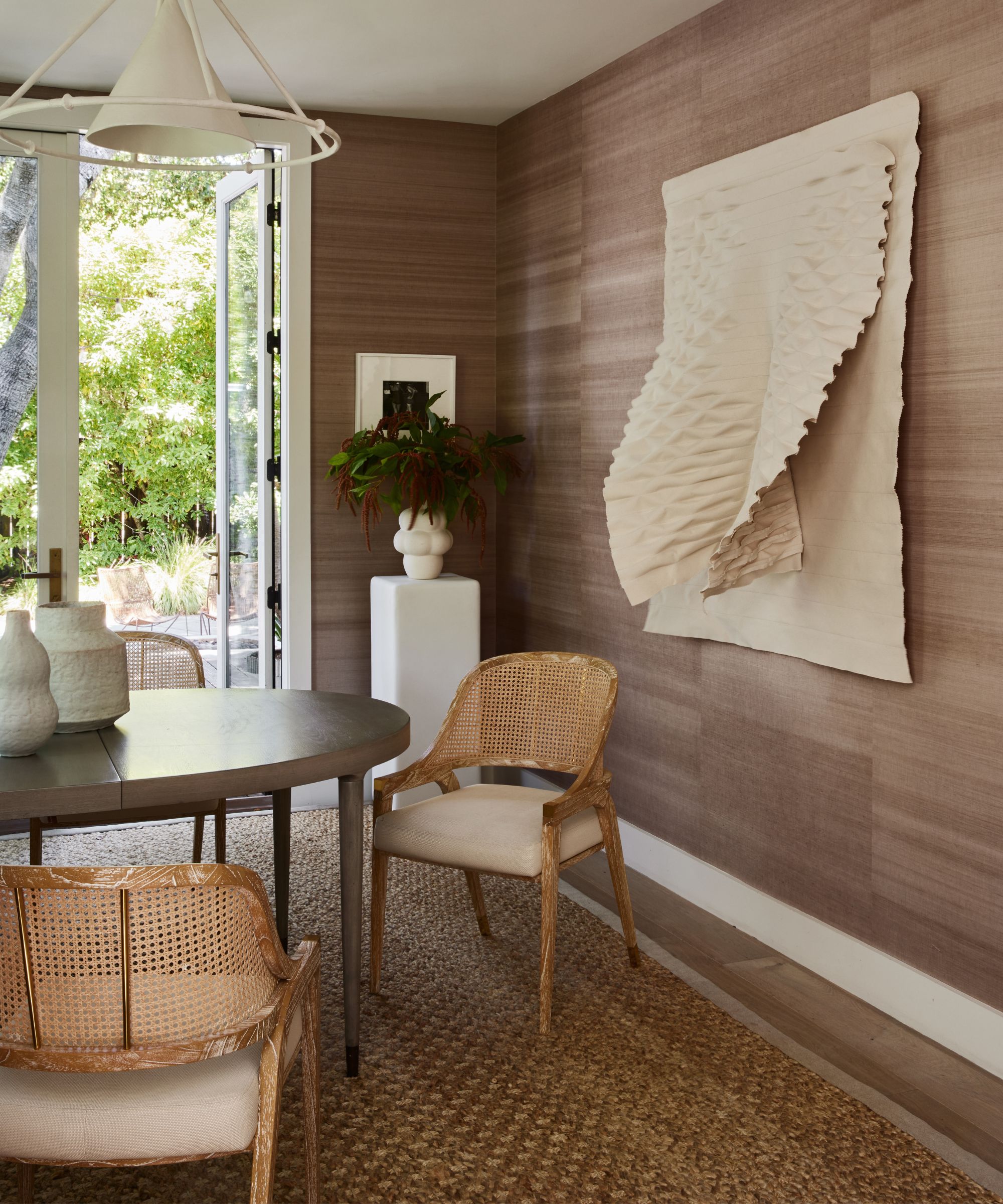 Neutral dining room with textured brown wallpaper and white textured hanging sculpture on wall, round wooden dining table with light rattan chairs