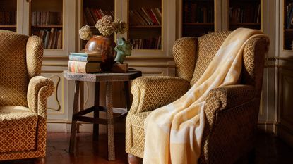 Linda Ring Fruttato wool yellow blanket draped on ahigh back arm chair with enclosed bookshelves behind