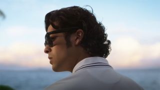 Still from the movie Blue Beetle (2023). Side profile of Jamie Reyes. He is wearing black sunglasses, a white polo shirt, with short, dark, curly hair. The ocean is in the background.