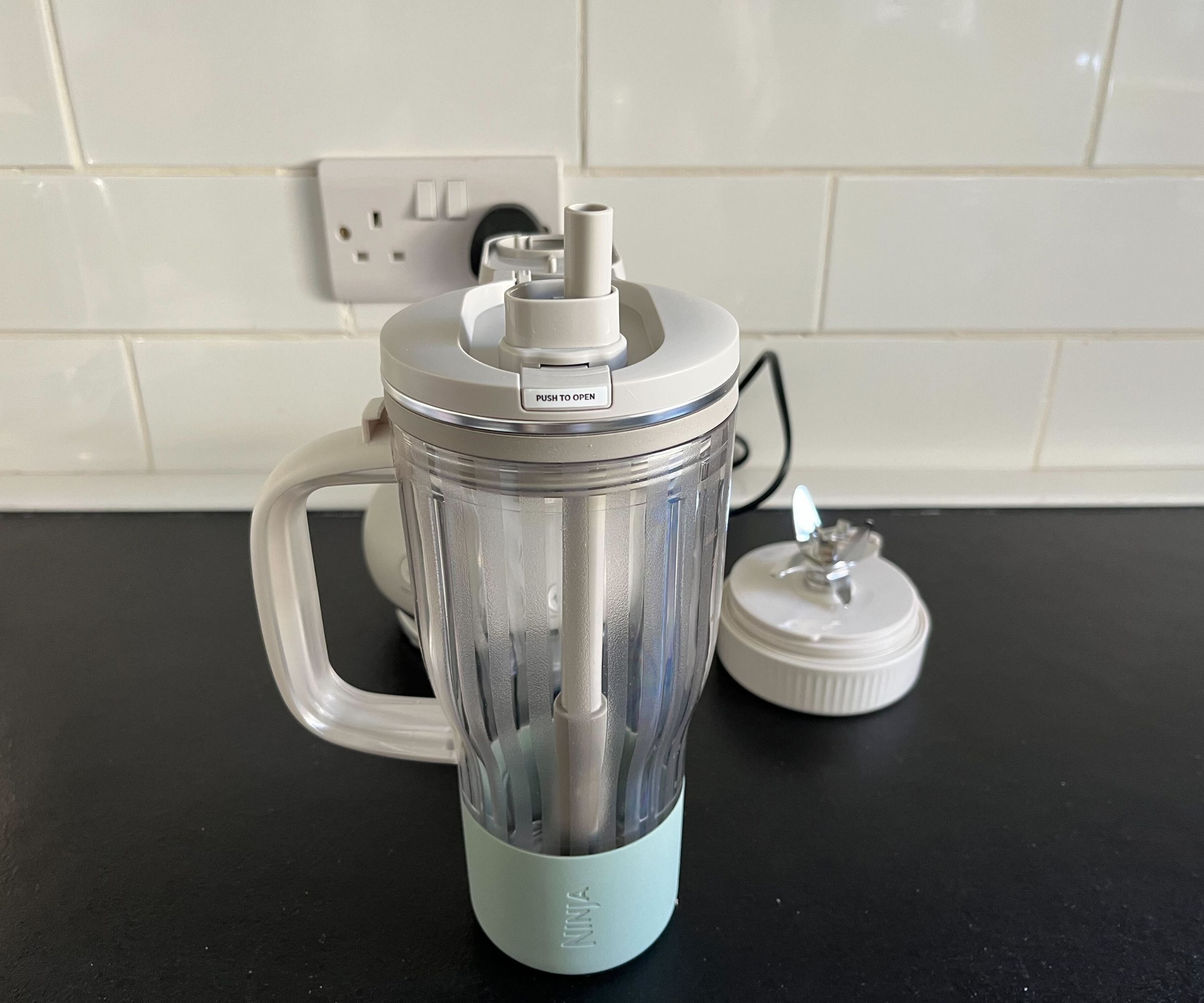 A clear plastic tumbler cup with a blue base and large grey handle, the lid is open and a straw is inside.