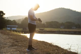 A man stood on the edge of a lake wearing shorts and a T-shirt looking at his watch