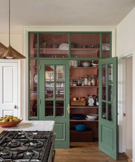 Kitchen with a built-in pantry in the corner, fitted with glass doors painted green. Inside, the pantry is painted pale pink, and shelves are filled with baking goods, ceramics, and kitchenware.