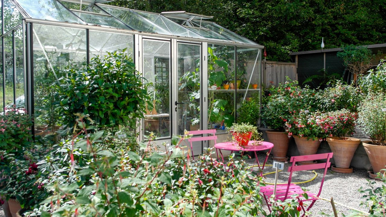 A close up of a flower garden with a greenhouse and pink table/seats 