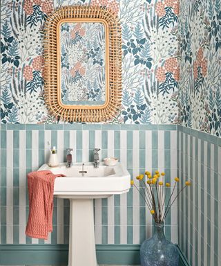 A bathroom with blue floral wallpaper, green and white vertically stacked tiles, a white sink, and a rattan mirror
