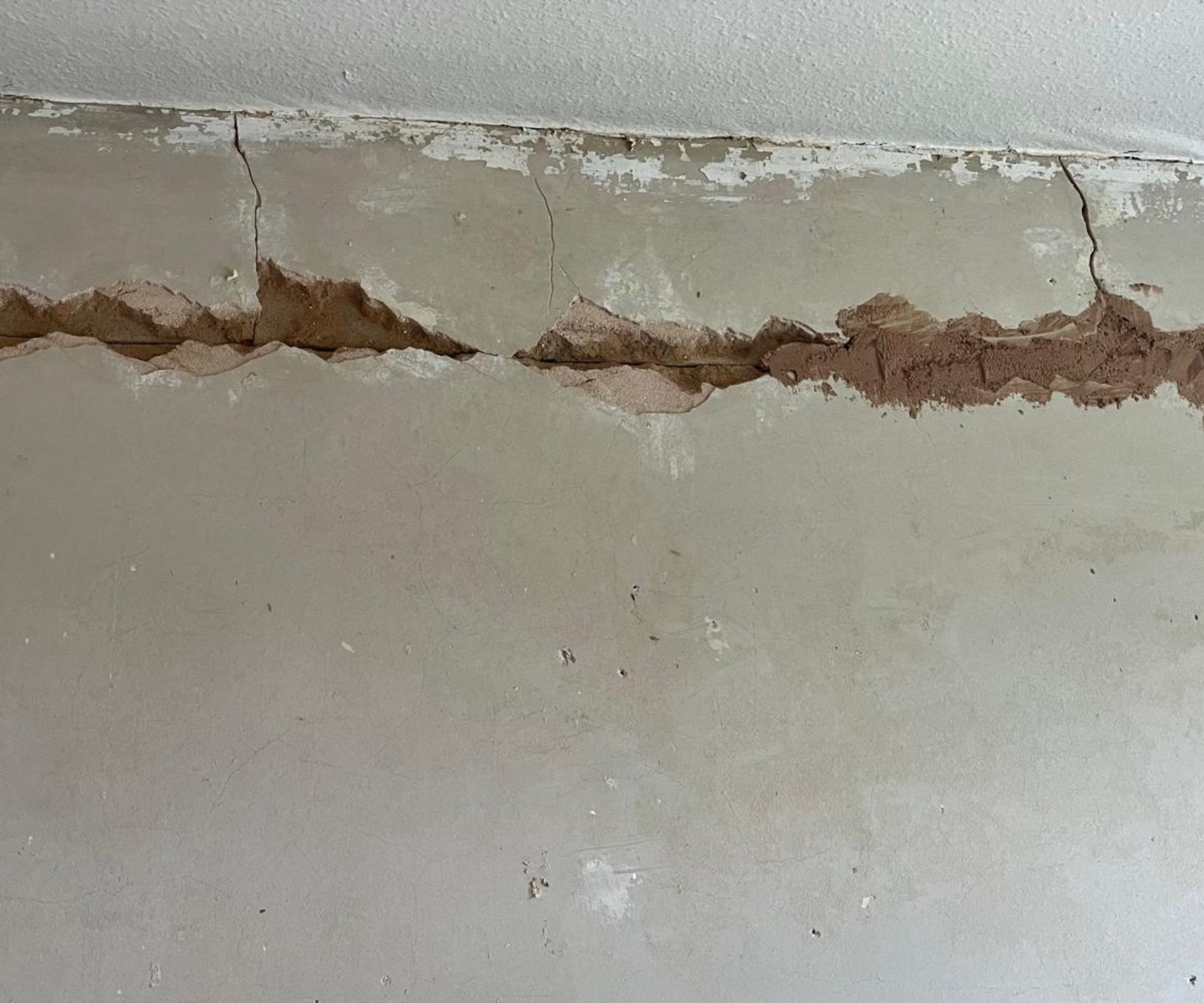 A close-up of a wall-ceiling junction showing significant plaster damage &amp;mdash; a long horizontal crack with crumbling, broken plaster exposing the brown render beneath.