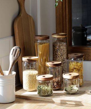 A light wooden kitchen counter with six glass jars filled with pasta, rice, and grains, white a white utensil jar next to them with wooden spatulas and a large dark brown wooden chopping board behind it