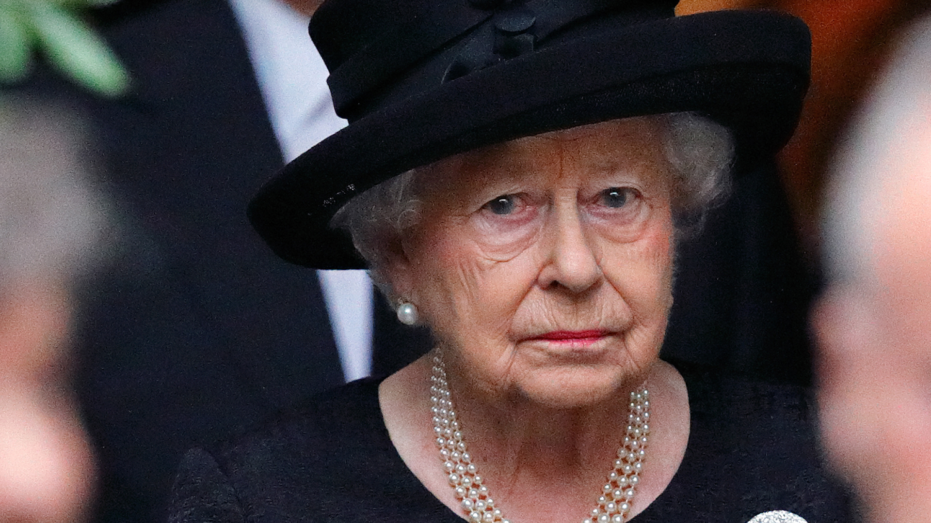 Queen Elizabeth II wears a black dress and matching hat and looks very sad while wearing a string of pearls