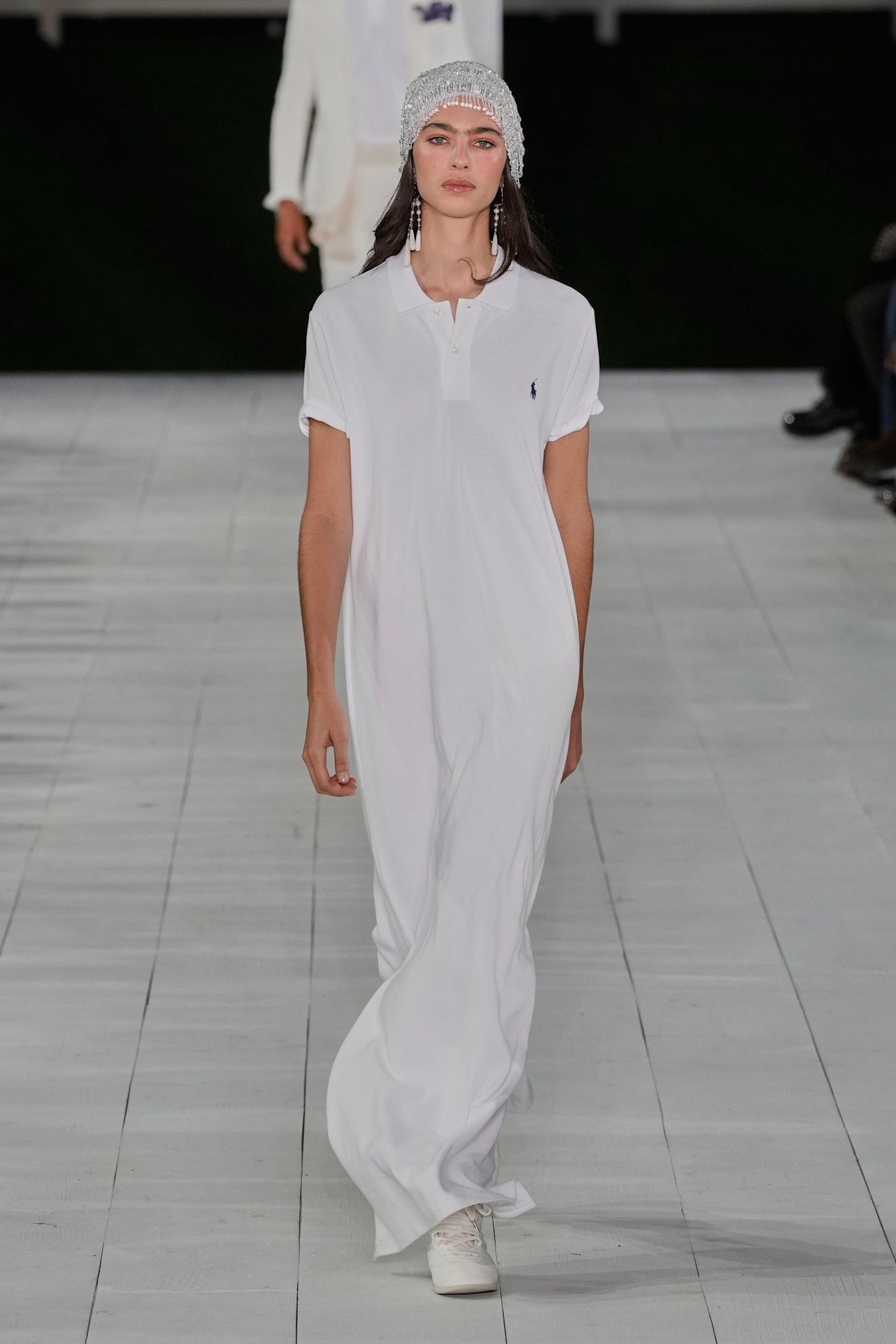 A model at the S/S 25 Ralph Lauren show wearing a white polo T-shirt dress with a beaded cap.