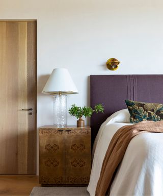 neutral bedroom with a linen purple headboard, white linen bedding, a tapestry throw pillow and a vintage style burl wood nightstand