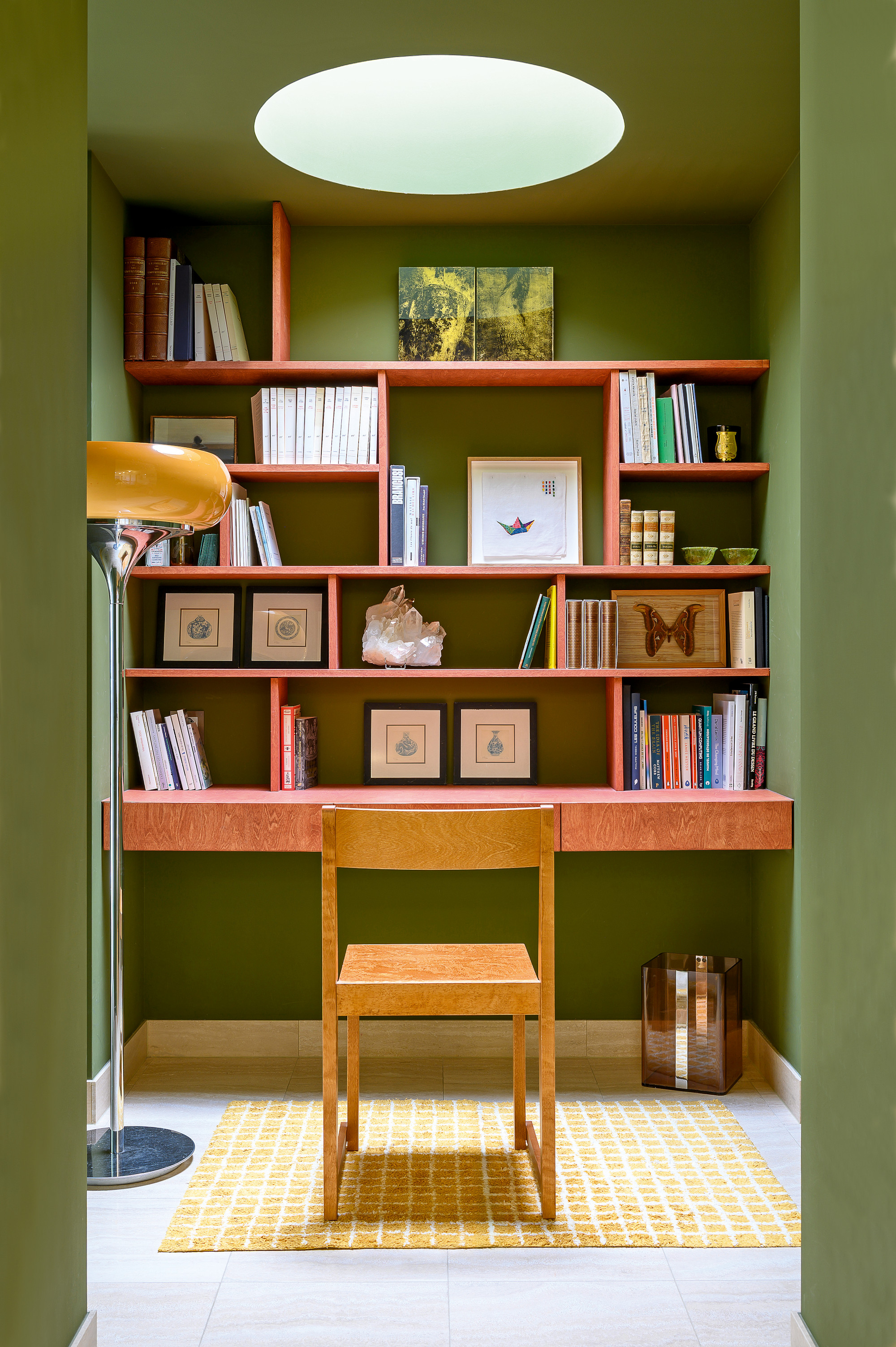 Office area with matcha green walls, natural light well, built-in irregular shelving in cherry wood, wood chair, yellow gridded rug and yellow and chrome floor lamp