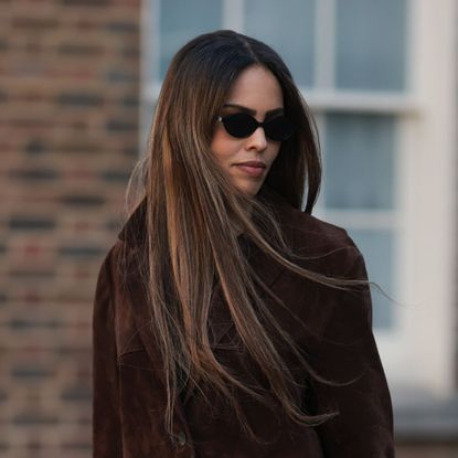 A woman walks down a London street wearing a brown suede jacket, sunglasses with beautiful glossy and healthy hair - best leave-in conditioner