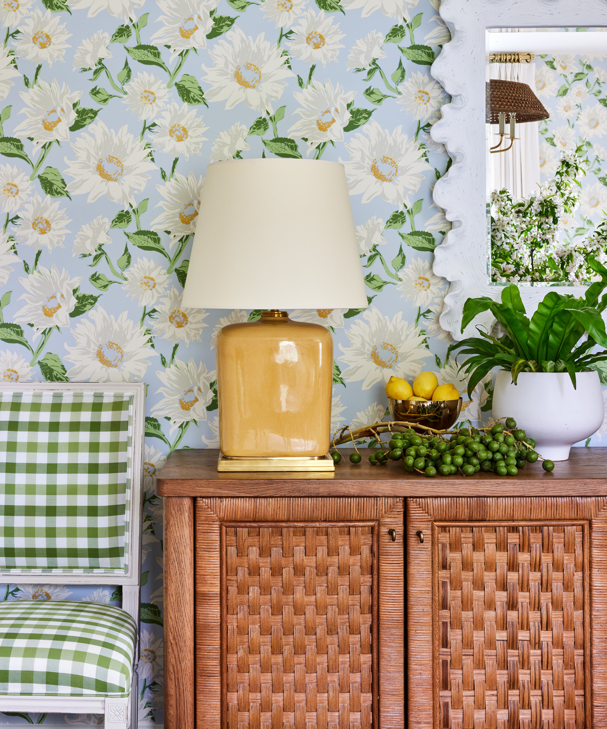 a rattan cabinet, a yellow lamp, a plant in a white pot, a green and white checker printed chair, and wallpaper with floral details