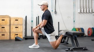 Andy Fata-Chan demonstrates the couch stretch using a weights bench in a gym. The top of his rear foot is resting on the bench, his rear knee is on the floor and his front foot is flat on the floor in front of him with his knee bent and above the ankle. Fata-Chan is wearing white cap, shorts and trainers, and a black T-shirt.