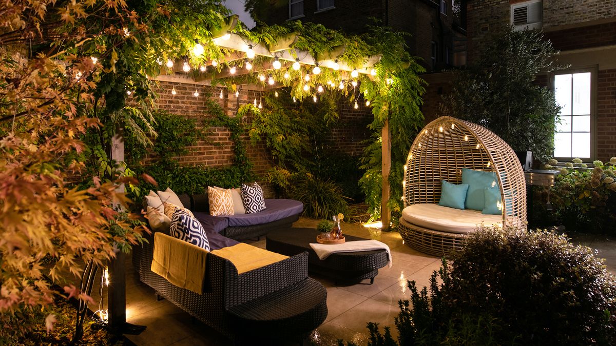 A well lit garden seating area with festoon lights in a pergola as well as lights around a rattan day bed