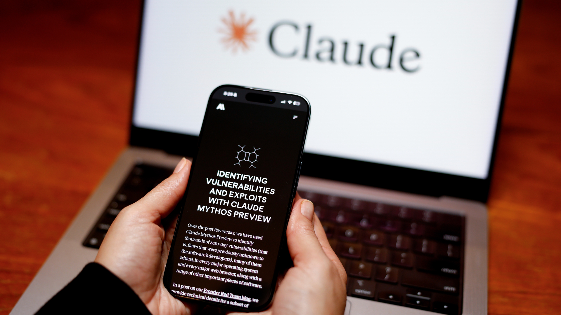 A smartphone has a black screen with the words: "Identifying vulnerabilities and exploits with Claude Mythos review" in front of a laptop with a white screen with the word "Claude" on it.