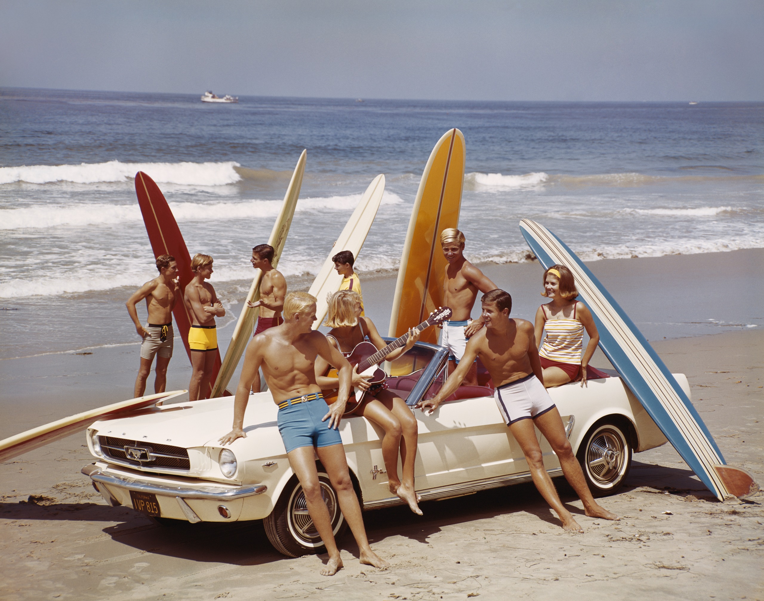 A group of blonde, tanned friends with surfboards surrounding a Ford Mustang on a California beach