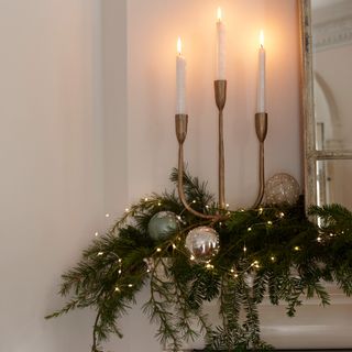 A fireplace mantelpiece decorated with a garland with string lights and metallic baubles and a candelabra with lit candles